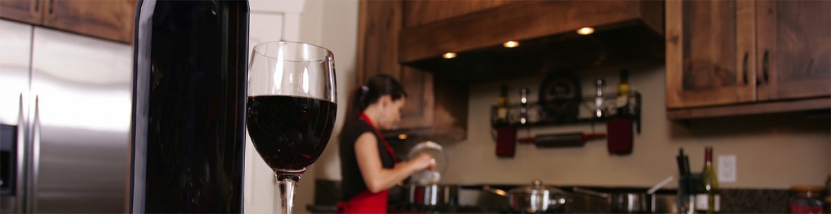 Verre de vin rouge et bouteille au premier plan d'une cuisine où une femme en tablier nettoie du matériel