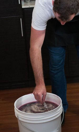 Un homme penché sur un seau de fermentation blanc brasse le moût violet et mousseux avec une cuillère