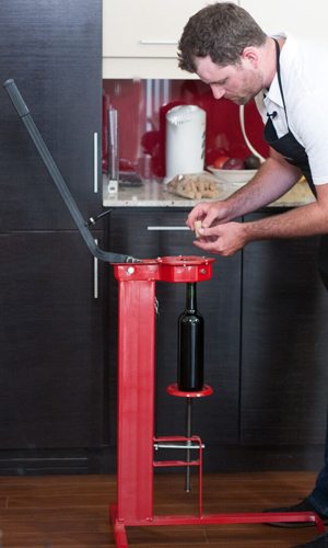 Une boucheuse sur pied rouge tenant une bouteille de vin, pendant qu'un vinificateur saisit un bouchon