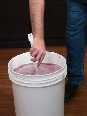 Un vinificateur brasse le moût rose et mousseux dans un seau de fermentation primaire blanc avec une longue cuillère