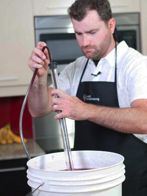 Un vinificateur plonge une canne de soutirage et un tuyau de siphon dans une cuve de moût rouge