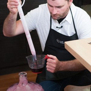Un vinificateur tient une longue pipette au-dessus d'une tasse à mesurer remplie de vin rouge, près d'une tourie