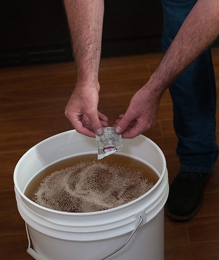 Mains qui saupoudrent un sachet de levure sèche sur le jus dans un seau de fermentation blanc