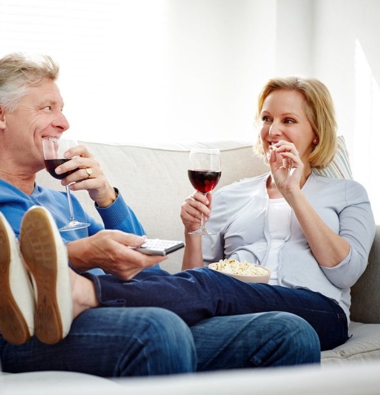 Happy mature couple relaxing together on sofa with popcorn and wine looking at each other while watching television at home